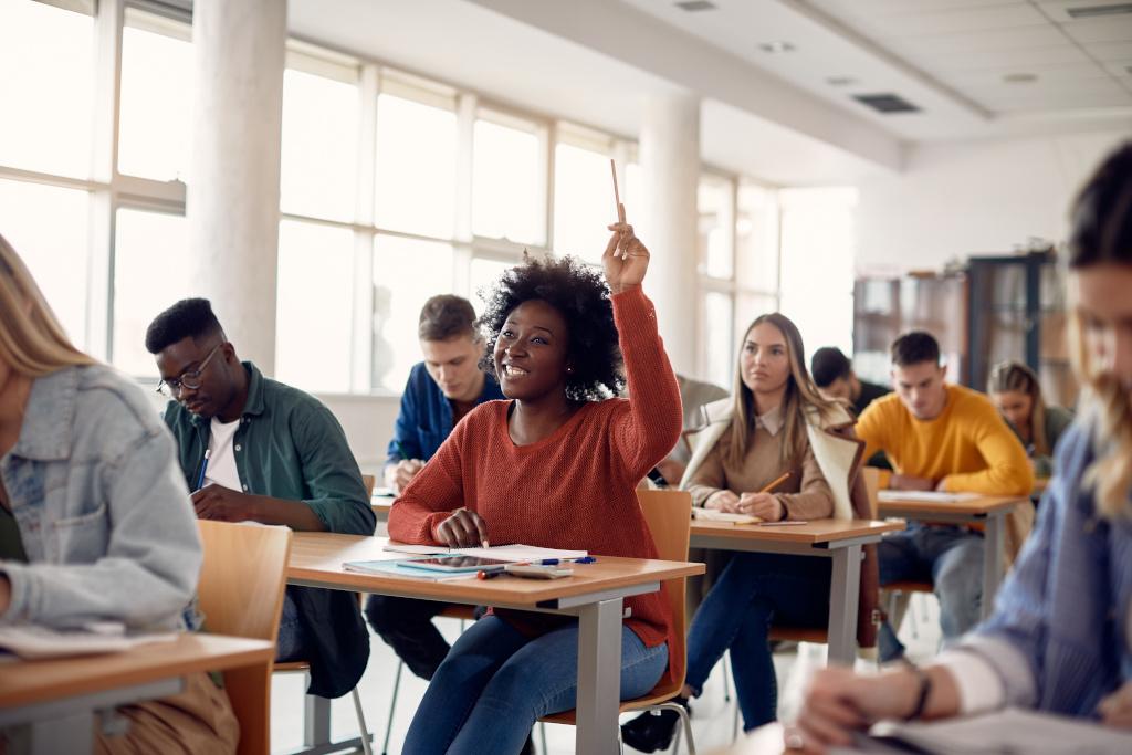 Students of a variety of races sit at tables in a white pillared classroom with a row of windows against wall. An African American female student, wearing a long-sleeved, ocher t-shirt, smiles and raises her hand.